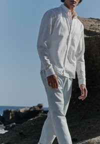 Hombre con camisa blanca y vaqueros claros caminando al aire libre cerca de un terreno rocoso bajo un cielo azul claro.