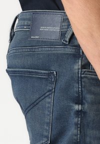 Jean en denim bleu foncé présentant une patch en cuir de marque à la taille, des poches latérales et un léger délavage sur la surface du tissu.