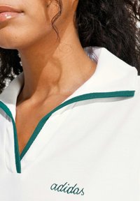 Biela polokošeľa s zelenými detailmi a golierom. Na hrudi je vyšívané logo "adidas" a je vyrobená z textúrovaného materiálu.