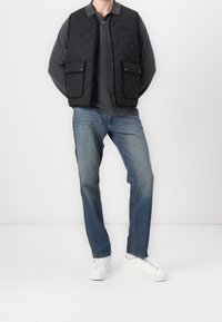 Černá prošívaná vesta se dvěma předními kapsami, nošená přes šedé tričko s dlouhým rukávem, v kombinaci s modrými džínami a bílými teniskami.