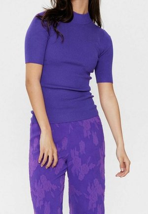 Kvinde iført en tætsiddende, ribstrikket lilla kortærmet top med højtaljede lilla bukser med blomsterprint, stående mod en ensartet lys baggrund.