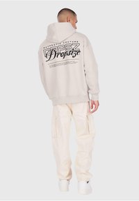 Jasno-beżowa bluza z kapturem z dużym czarnym napisem na plecach, połączona z kremowymi spodniami cargo i białymi trampkami. Prosty, luźny krój.