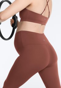 Femme en brassière de sport marron et leggings tenant un cercle de Pilates noir, vue de dos et de côté sur fond blanc.