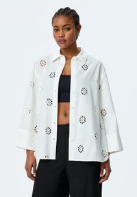 White cotton shirt with floral cut-out details, button closure, and oversized fit. Paired with a black top and textured black pants.