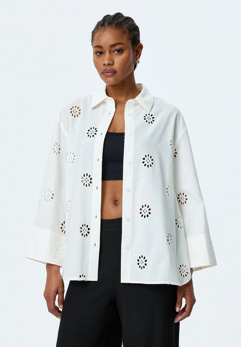White cotton shirt with floral cut-out details, button closure, and oversized fit. Paired with a black top and textured black pants.