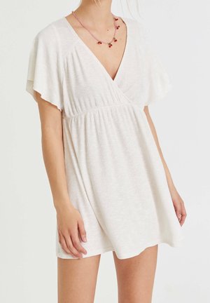 Femme portant une courte robe blanche côtelée avec des manches volantées et un col en V, accessoirisée d'un collier rose orné de petites fleurs rouges.