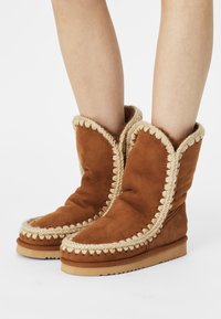 Bottines en suède marron avec une texture douce, dotées d'un galon tressé crème décoratif le long des bords et d'une semelle plate.