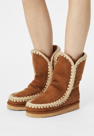 Bottines en suède marron avec une texture douce, dotées d'un galon tressé crème décoratif le long des bords et d'une semelle plate.