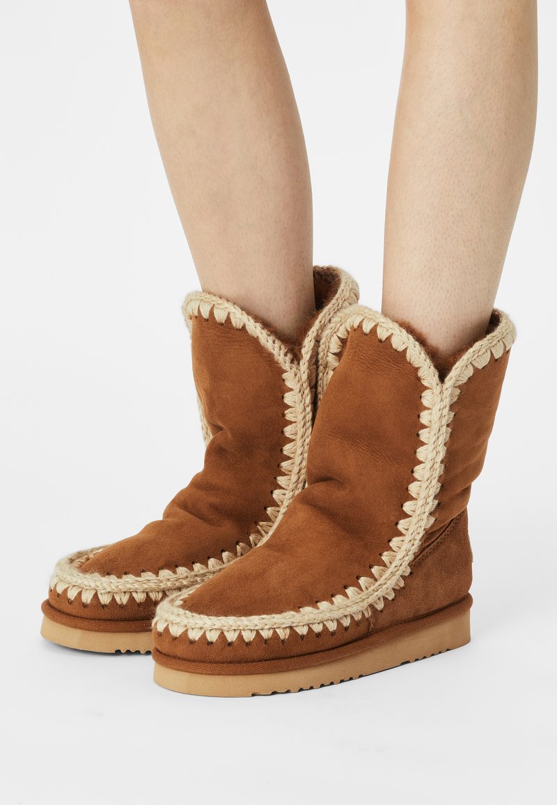 Bottines en suède marron avec une texture douce, dotées d'un galon tressé crème décoratif le long des bords et d'une semelle plate.