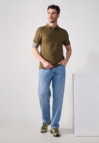 Polo verde oliva, vaqueros rectos azules y zapatillas verdes con patrón. Ajuste casual y tejido suave con un cuello clásico.