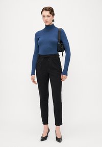 Top de cuello alto de manga larga azul combinado con pantalones negros ajustados que tienen una cintura elástica. Bolso negro y tacones de punta negra.