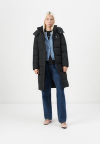 Svart pufferjakke med hette og quiltet design, lagd over en blå cardigan, kombinert med vide jeans og lys rosa sko.