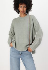 Grüner Strickpullover mit ribbeligem Saum und Bündchen, der einen lockeren Schnitt und überschnittene Schultern aufweist, getragen mit hellblauen Jeans.