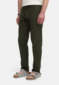 Olivenfarvede teksturerede bukser med snoreliv, der har læg og sidesømme, parret med grå sandaler med justerbare stropper.