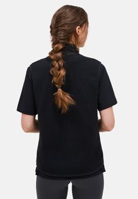 Schwarze Fleece-Weste mit einer strukturierten, weichen Oberfläche; ärmelloses Design; über einem schwarzen Kurzarmshirt getragen; lange, geflochtene Haare sichtbar.