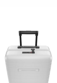 Valise rigide blanche avec une poignée noire rétractable, un verrou à combinaison et des rainures horizontales sur la surface pour la texture.