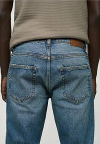 Rückansicht einer Person, die eine hellblaue Jeans und ein beigefarbenes, strukturiertes Oberteil trägt. Die Rückentaschen und die Gürtelschlaufen sind sichtbar.