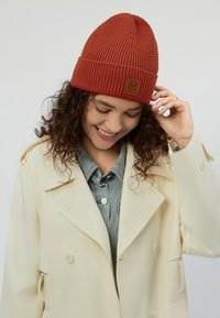 Rustfarvet ribstrikket hue med opsmøg og en brun logoemblem. Båret af en person iført en lys beige frakke over en denimskjorte.