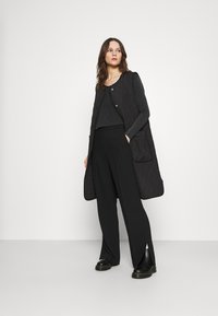 Veste longue en cuir matelassé noir avec des boutons dorés, portée sur un haut sombre et un pantalon large noir avec des fentes. Des chaussures noires épaisses complètent le look.