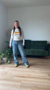 Roupa casual com uma camisa de mangas longas branca com texto em vermelho e amarelo, jeans largos azuis e sapatos amarelos, em uma sala iluminada com um sofá verde.