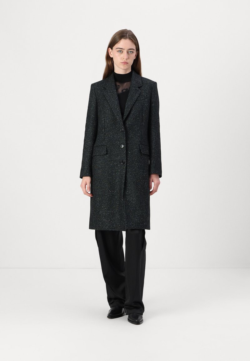 Manteau long noir texturé avec un design ajusté, fermeture à un bouton et deux poches avant, porté sur un haut et un pantalon noirs.