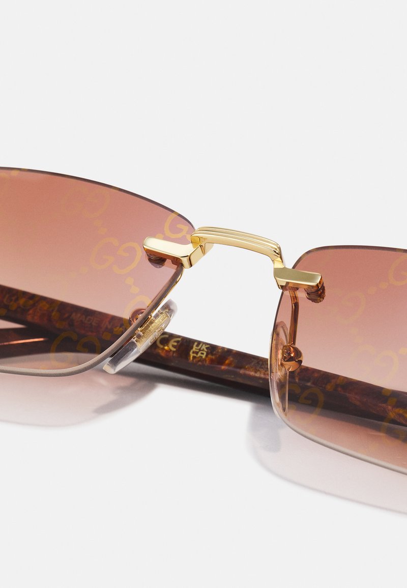 Lunettes de soleil dégradées brunes avec des accents dorés, branches épaisses en écaille de tortue et détails de logo sur les verres. Matériaux en plastique et métal lisses.