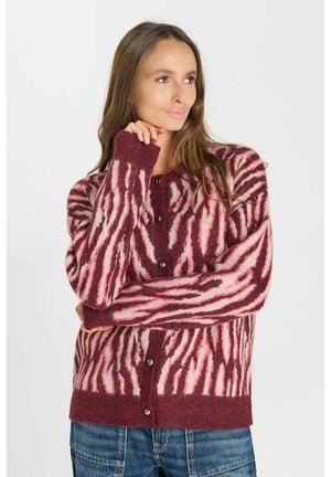 Vrouw met lang bruin haar die een rood- en roze patroon gebreid vest met knopen en een blauwe spijkerbroek draagt, poseert met één hand bij haar gezicht.