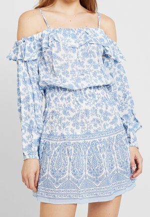 Kvinde iført en hvid off-shoulder kjole med blå blomsterprint, flæset halsudskæring og lange ærmer med elastiske manchetter.