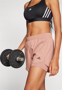 Crna sportska grudnjak s bijelim detaljima, uparen s ružičastim šortsevima s elastičnom trakom i Adidas logom. Osoba drži bučicu.