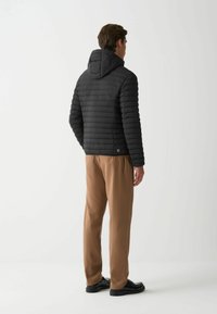 Chaqueta de plumón negra con patrón de acolchado horizontal, capucha adjunta y detalle de logo. Lleva puesta con pantalones marrones y zapatos negros.