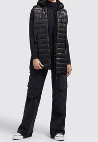 Czarna pikowana kamizelka z przeszyciami, kapturem, zapinanymi na zamek kieszeniami i długim krojem. W zestawieniu z ciemnymi bojówkami i białymi tenisówkami.