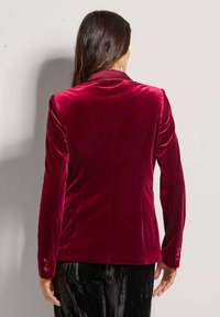 Fluwelen bordeauxkleurige blazer met een getailleerde pasvorm, spitse revers en drie knopen op elke manchette. Glad textuur en gestructureerd ontwerp.