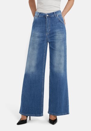 Vrouw draagt een blauwe spijkerbroek met hoge taille en wijde pijpen met vervaagde vlekken, gecombineerd met zwarte spitse hakken en een wit ingestopt topje.