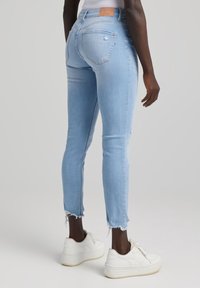 Vaqueros ajustados de mezclilla azul claro con dobladillos deshilachados, que presentan una silueta ajustada y dos bolsillos traseros. Combinados con zapatillas blancas.