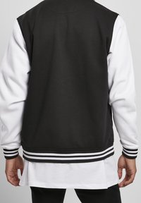 Veste universitaire noire et blanche avec une texture lisse, des poignets rayés et une coupe décontractée. Le dos présente un design noir uni.