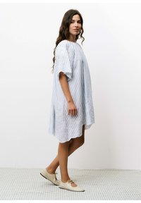 Blauw-wit gestreepte jurk met korte mouwen, voorzien van kantdetails aan de onderkant. G gedragen met lichtgekleurde, platte schoenen.