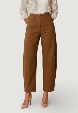 Donna che indossa pantaloni marroni a gamba larga e vita alta, camicia a righe con bottoni e maniche arrotolate e scarpe con tacco nude.