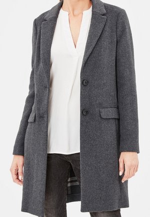 Mujer con un abrigo de lana gris oscuro hasta la rodilla, sobre una blusa blanca de escote en V y pantalones oscuros, de pie frente a un fondo liso.