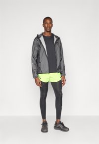 Giacca leggera nera con interno a fantasia, camicia nera, pantaloni corti verdi neon, leggings neri e scarpe da corsa nere.