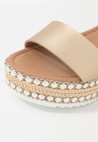 Beige Sandale mit offenem Zehenbereich, dicker Riemen, geflochtener Espadrille-Sohle und zwei Reihen runder, metallischer Nieten am Rand.