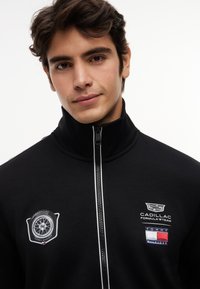 Mladý muž oblečený v čiernej bunde na zips s logami tímu Cadillac Formula 1 a Tommy Hilfiger na hrudi na bielom pozadí.