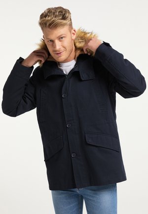 Homme blond portant une veste d'hiver bleu foncé avec une capuche en fourrure, ajustant la capuche en se tenant devant un fond clair uni.