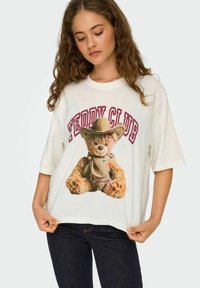 Biała, oversize'owa koszulka z nadrukiem misia w kowbojskim kapeluszu i bandanie, z napisem "Teddy Club" powyżej w burgundowym kolorze.