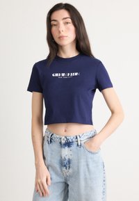 Tamsiai mėlynos spalvos trumpas medvilninis marškinėliai su trumpomis rankovėmis ir apvaliu iškirpimu. Pasipuošę balto logotipo spausdinimu "CALVIN KLEIN NEW YORK CITY."