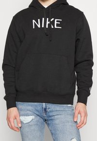 Svart bomullshoodie med frontficka, dragsko för huvan och vit "NIKE"-logotyp. Bärs över ljusblå slitna jeans.