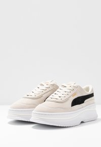 Beige mokkasiner med sorte detaljer på siden, hvit tykk såle og kontrasterende beige lisser. Flatt tekstur og rund tå-design.