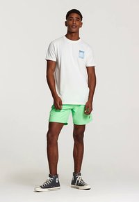 Wit katoenen T-shirt met een klein blauw grafisch zakje en korte mouwen, gecombineerd met felgroene korte broek en grijze hoge sneakers.