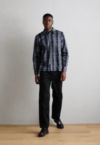 Camisa de manga longa preta com um padrão abstrato cinza, combinada com calças pretas e sapatos pretos, em um fundo neutro.