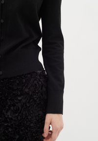 Cardigan nero con chiusura a bottoni, realizzato in tessuto morbido. Abbinato a una gonna nera floreale con texture, che mette in mostra dettagli a strati.