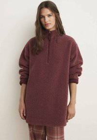 Sweater de forro burdeos con cuello alto y media cremallera, que presenta mangas XL y una superficie texturizada. Combinado con pantalones de cuadros.